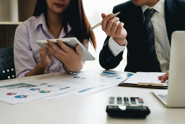 Two business professionals sitting at a desk analyzing documents and data on a digital tablet.